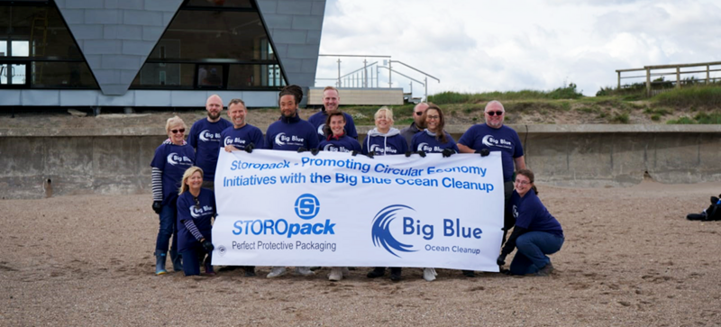 Varias personas posan para una foto de grupo en la playa sosteniendo una pancarta de Big Blue Ocean Cleanup